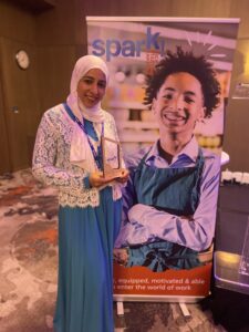 Ruhina with the Spark Award in front of Award poster.