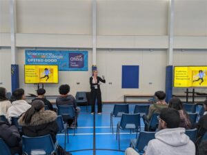 Group of students in sitting in a sports hall listening to female speaker.