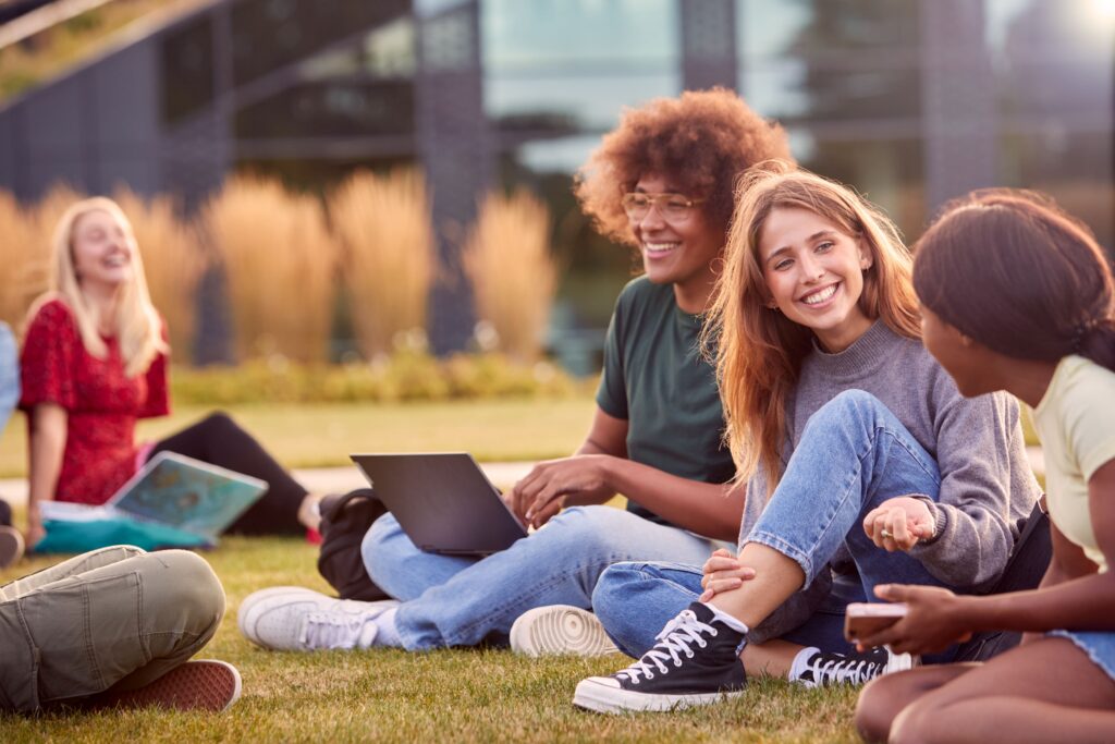 Stanmore College - Group of students on gras gathering around with laptops smiling.